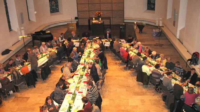 Große Gruppe von Menschen sitzt an langen Tischen in einem Raum und isst.
