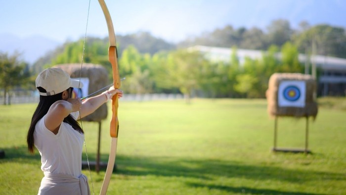Eine Person übt im Freien Bogenschießen und zielt mit einem Bogen auf eine Zielscheibe auf einem grasbewachsenen Feld.