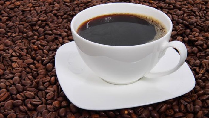 En hvid kaffekop fyldt med sort kaffe står på en underkop omgivet af kaffebønner.