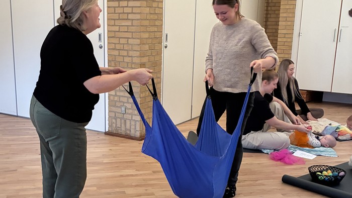 To kvinder justerer en blå hængekøje i et rum, hvor en anden kvinde og et barn er i nærheden.