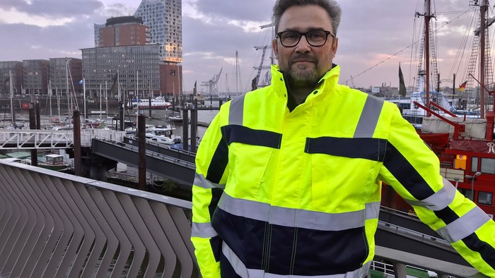 En mand i højlysende arbejdstøj står ved en havn med moderne bybygninger i baggrunden.