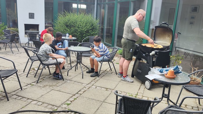 En mand stoker grill i en udendørs spiseområde, hvor andre gæster sidder og spiser.