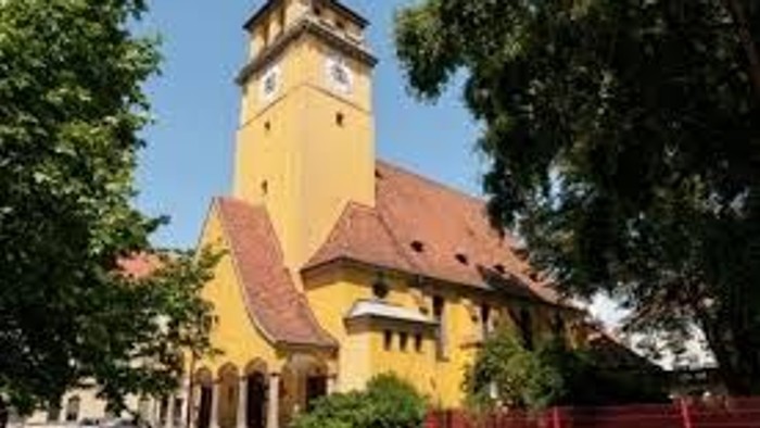 Bezaubernde gelbe Kirche mit hohem Uhrturm und rotem Ziegeldach, eingebettet zwischen grünen Bäumen.