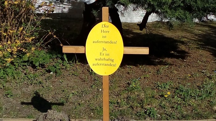 Ein hölzernes Kreuz mit einem gelben Schild und Muscheln am Fuß in einer gestalteten Grünanlage.