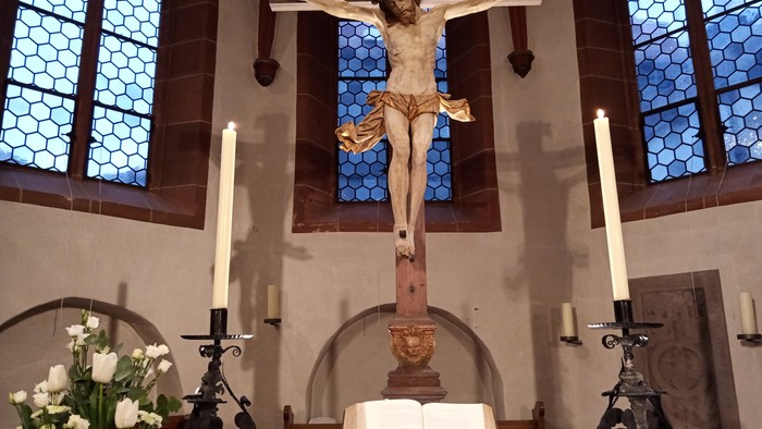 Das Bild zeigt eine Kircheninnenansicht mit einem Kruzifix und Kerzen.