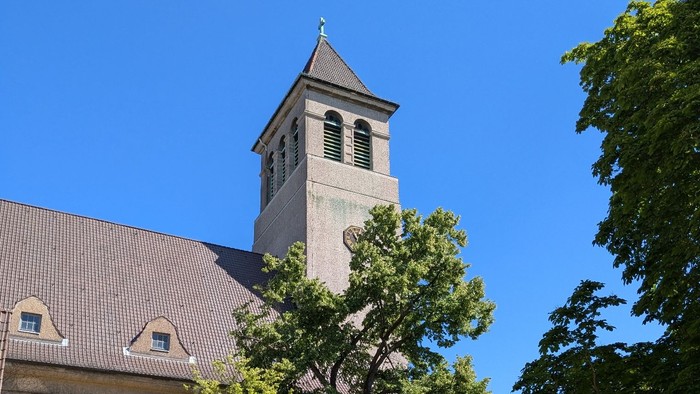 Kirchturm mit Uhr und Kreuz, umgeben von Bäumen und Gebäuden