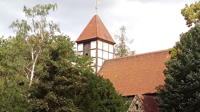 Das Bild zeigt eine Kirche mit rotem Dach und einem Turm, umgeben von Bäumen.