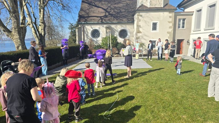 En livlig udendørs sammenkomst med børn og voksne nær en kirke, hvor de holder farverige paraplyer og legetøj.
