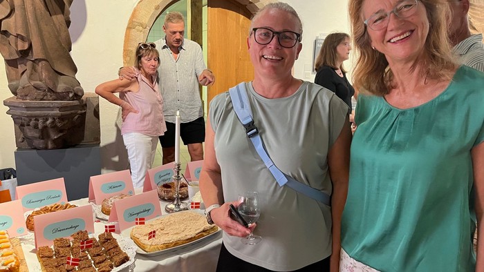 To glade kvinder står ved et madbord til en social sammenkomst med andre i baggrunden.