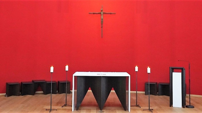 Moderne Altarraum-Gestaltung mit einer leuchtend roten Rückwand, vor der ein filigranes Metall-Kruzifix hängt. Im Zentrum steht ein dunkler, geometrischer Altar mit weißer Platte, flankiert von zwei schlanken Kerzenständern.