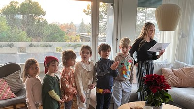 Børn står opmærksomt, mens en voksen læser en bog i en hyggelig, veloplyst stue.