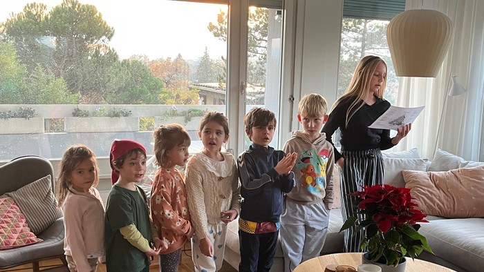 Børn står opmærksomt, mens en voksen læser en bog i en hyggelig, veloplyst stue.