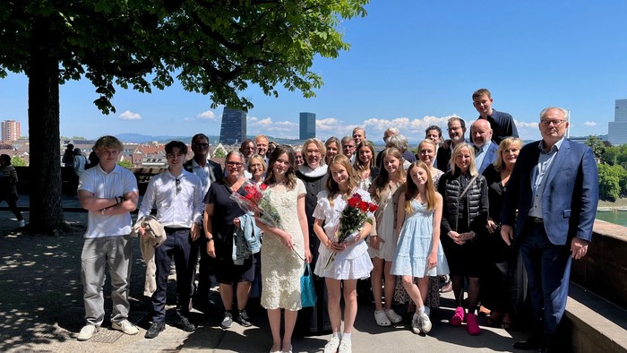 Gruppefejring med dimittender, der holder blomster og poser udendørs nær et naturskønt bylandskab under solskinsvejr.