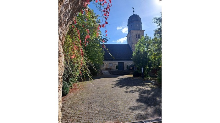 Kirche mit hohem Turm und roten Blumen an einem baumgesäumten Weg.