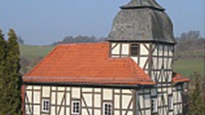 Traditionelles Fachwerkhaus mit rotem Dach und Turm, umgeben von ländlicher Umgebung.