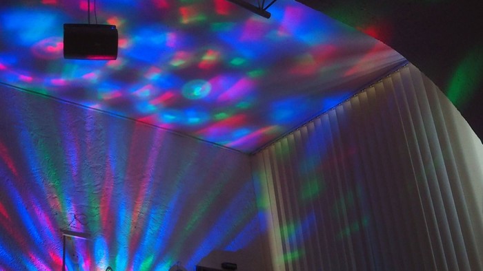 Lebendige Laserlichter erzeugen eine farbenfrohe Deckenprojektion in einem schwach beleuchteten Raum, in dem sich jemand auf einer Couch entspannt.