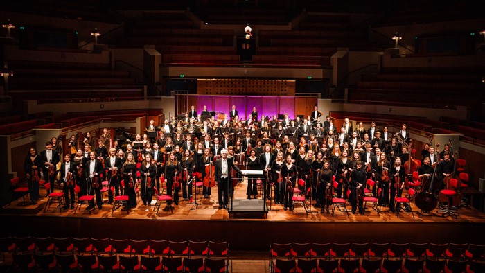 Et stort orkester optræder på scenen i en stor, svagt oplyst koncertsal.