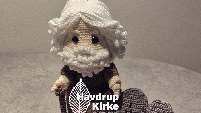 Hæklet figur, der holder et farverigt kors med gravsten og hat, som repræsenterer et religiøst tema.