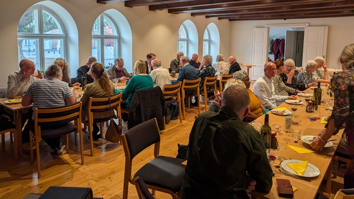 Gruppemiddag i et rummeligt lokale med buede vinduer, hvor folk snakker og nyder måltider sammen.