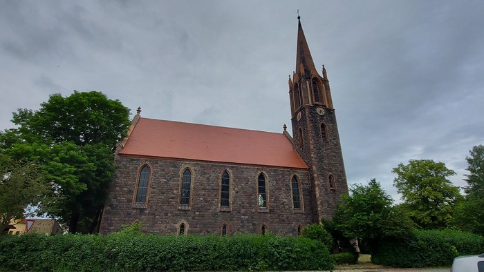 Ein Kirchengebäude mit hohem Turm und rotem Dach, umgeben von Bäumen und Büschen.