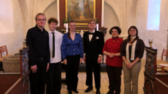 Gruppen står i en kirke og holder et stort religiøst ikon, mens de poser nær kirkebænke og et dekoreret alter.