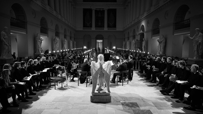 En sort-hvid fotografi af en modeforestilling i et stort hallen med statuer og en stor publikum.