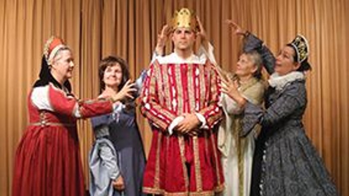 Auf einer Bühne vor einem geschlossenen, goldfarbenen Vorhang steht eine Person in der Mitte in einem auffälligen roten Gewand mit goldenen Verzierungen und einer Krone. Die Figur wirkt ernst und aufrecht, die Hände vor dem Körper verschränkt. Vier weiter