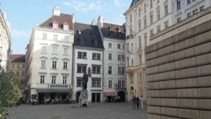Städtischer Platz, umgeben von historischen europäischen Gebäuden mit einer Statue und einer modernen Holzkonstruktion im Vordergrund.