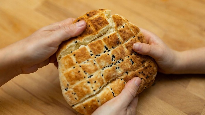 Zwei Personen halten gemeinsam ein rundes, goldbraun gebackenes Fladenbrot über einer Holzoberfläche. Das Brot ist in kleine Quadrate eingeritzt und mit schwarzen Samen bestreut. Die Hände greifen von gegenüberliegenden Seiten nach dem Brot und ziehen es 