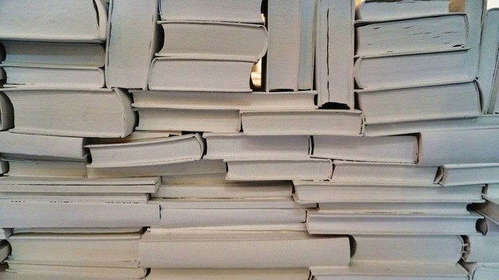 Das Bild zeigt einen dicht gestapelten Turm aus zahlreichen Büchern. Die Bücher liegen quer und längs übereinander, sodass ein unregelmäßiges, fast mauerartiges Muster entsteht. Sichtbar sind vor allem die hellen Buchschnitte und die unteren Kanten der Ei