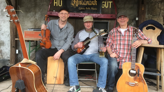 Drei erwachsene Männer sitzen nebeneinander auf einer kleinen Holzplattform vor einer rustikalen Stein- und Holzwand. Über ihnen hängt ein Schild mit der Aufschrift „Amarank – Celtic Folk Music“. Alle drei lächeln in die Kamera und halten Musikinstrumente