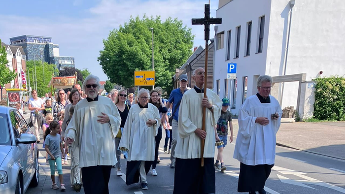 Eine Gruppe von Priestern in weißen Gewändern führt eine Prozession auf einer Stadtstraße an, begleitet von Gläubigen.