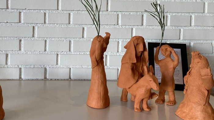 Håndlavede terrakotta-skulpturer af elefanter og menneskefigurer arrangeret på en lys overflade foran en murstensvæg.