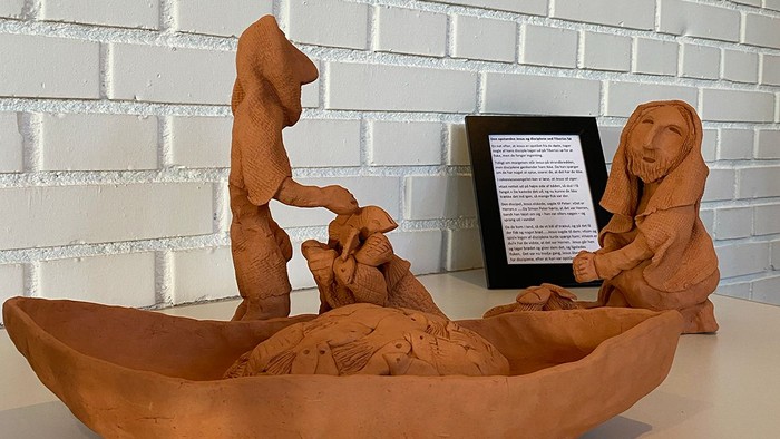 Lerfigur af en båd med figurer og en fritstående siddende figur ved siden af en plakette.