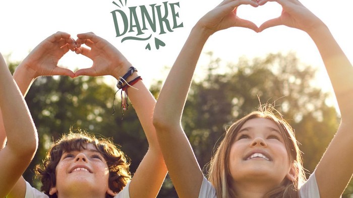 Zwei fröhliche Kinder formen mit ihren Armen Herzformen im Freien, lächeln in der Sonne und über ihnen steht „Danke“.