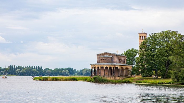 Ein großes Gebäude am Wasser mit Säulen und einem Turm, umgeben von Bäumen und einem klaren Himmel.