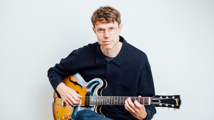 En person med briller spiller på en elguitar iført en afslappet, mørk sweater og jeans.