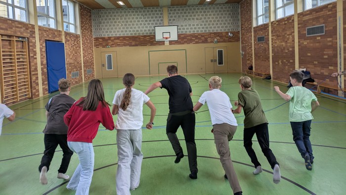 Gruppe von Menschen, die in einer Turnhalle mit grünem Boden und Backsteinwänden ein Kreisspiel spielen.