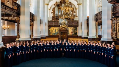 Et stort kor optræder i en prægtig kirke, siddende halvcirkelformet foran et udsmykket orgel og alter.