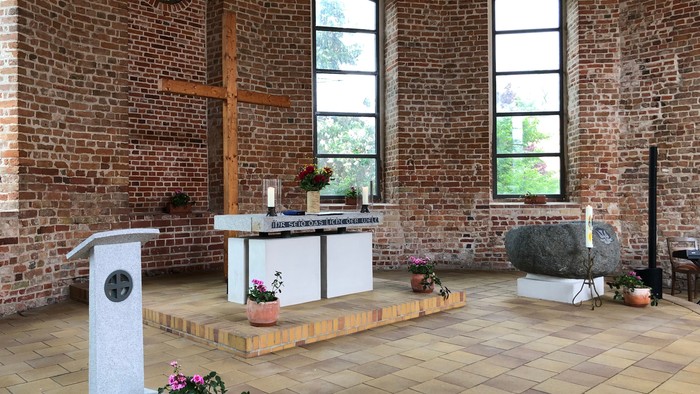 Ein ruhiger Kapelleninnenraum mit einem schlichten Altar, einer Kanzel und dekorativen Pflanzen vor Ziegelwänden und großen Fenstern.