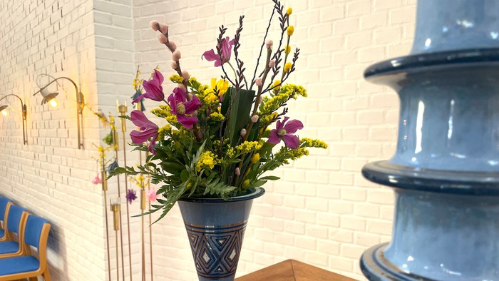 Farverigt blomsterarrangement i en dekorativ vase på et træbord ved siden af en blå stol.