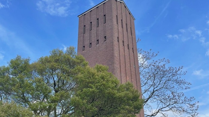 Turm mit Uhr und Kreuz vor blauem Himmel und Bäumen