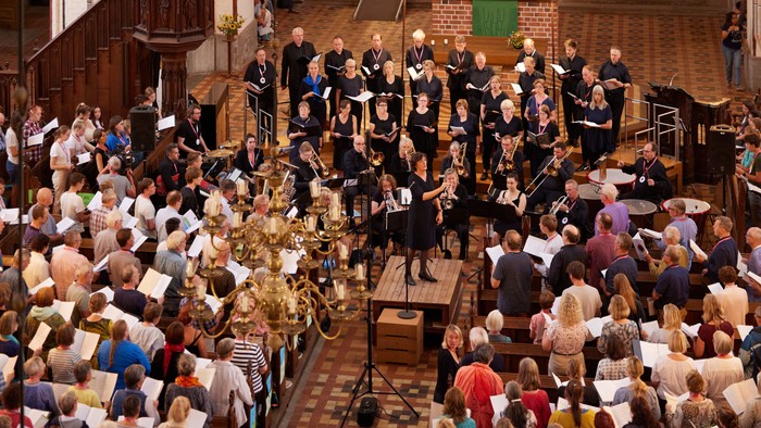 Ein großer Chor und ein Orchester treten gemeinsam in einer prächtigen, historischen Kirche auf, die mit aufmerksamen Zuhörern gefüllt ist.