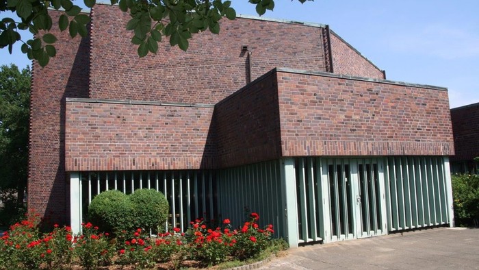 Das Bild zeigt ein modernes Ziegelgebäude mit grünen Fensterläden und blühenden Pflanzen davor.