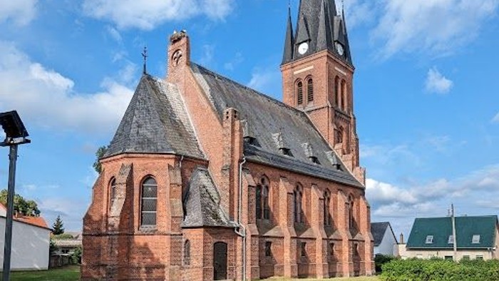 Historische Backsteinkirche mit hohem Turm, gelegen in einer grasbewachsenen, offenen Fläche unter klarem Himmel.