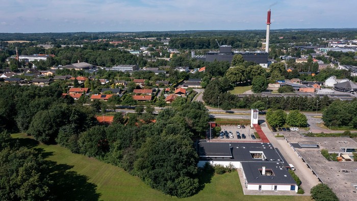 En stor industrikompleks med en høj røgkamin i en by omgivet af grønne områder.