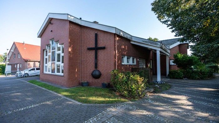 Ziegelkirche mit Kreuzsymbol an der Fassade, umgeben von Bäumen und anderen Gebäuden.