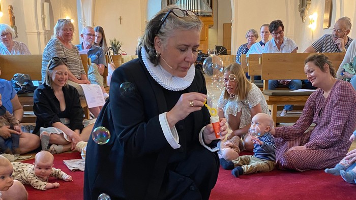 En kvinde i mørkt kapel let sig se med børn og forældre i kirke.