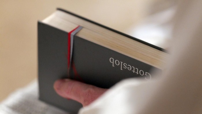 Eine Hand hält ein schwarzes Buch mit der Aufschrift „Gotteslob“, das mit einem roten Band gesichert ist.