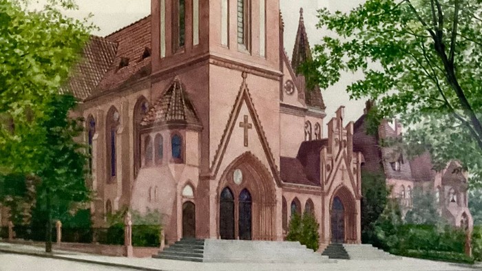 Gotische Kirche mit hohem Turm, eingerahmt von üppigen grünen Bäumen unter bewölktem Himmel.